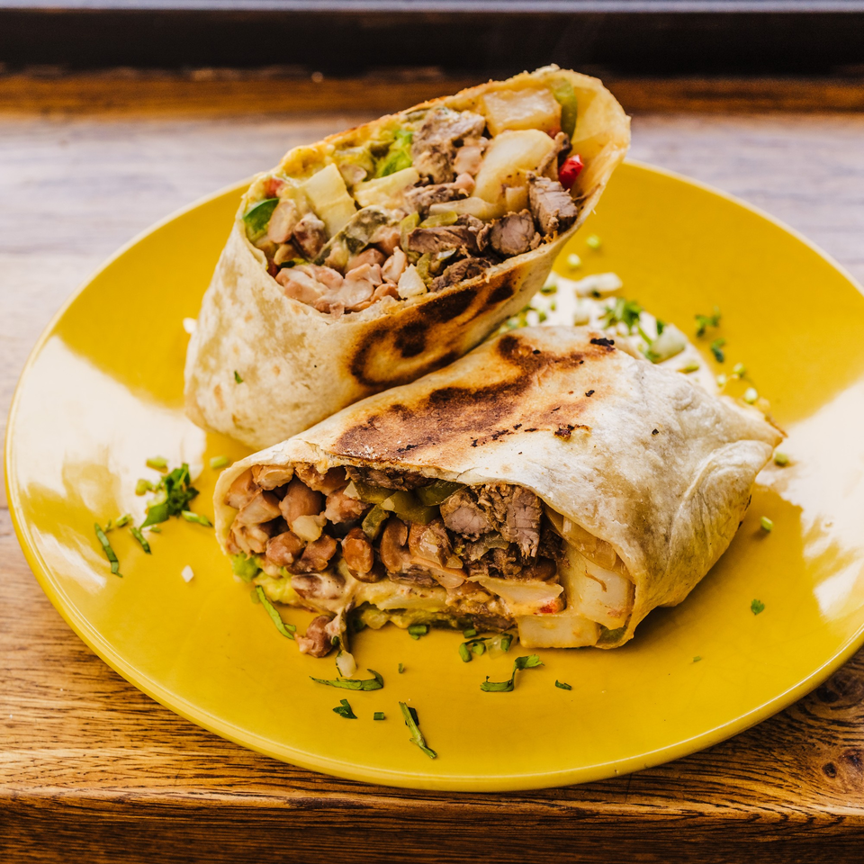 Two halves of a large, grilled burrito filled with meat, beans, and potatoes on a yellow plate.